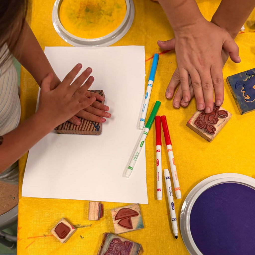 Hands press wooden stamps onto paper at a yellow table with colorful markers and ink pads.