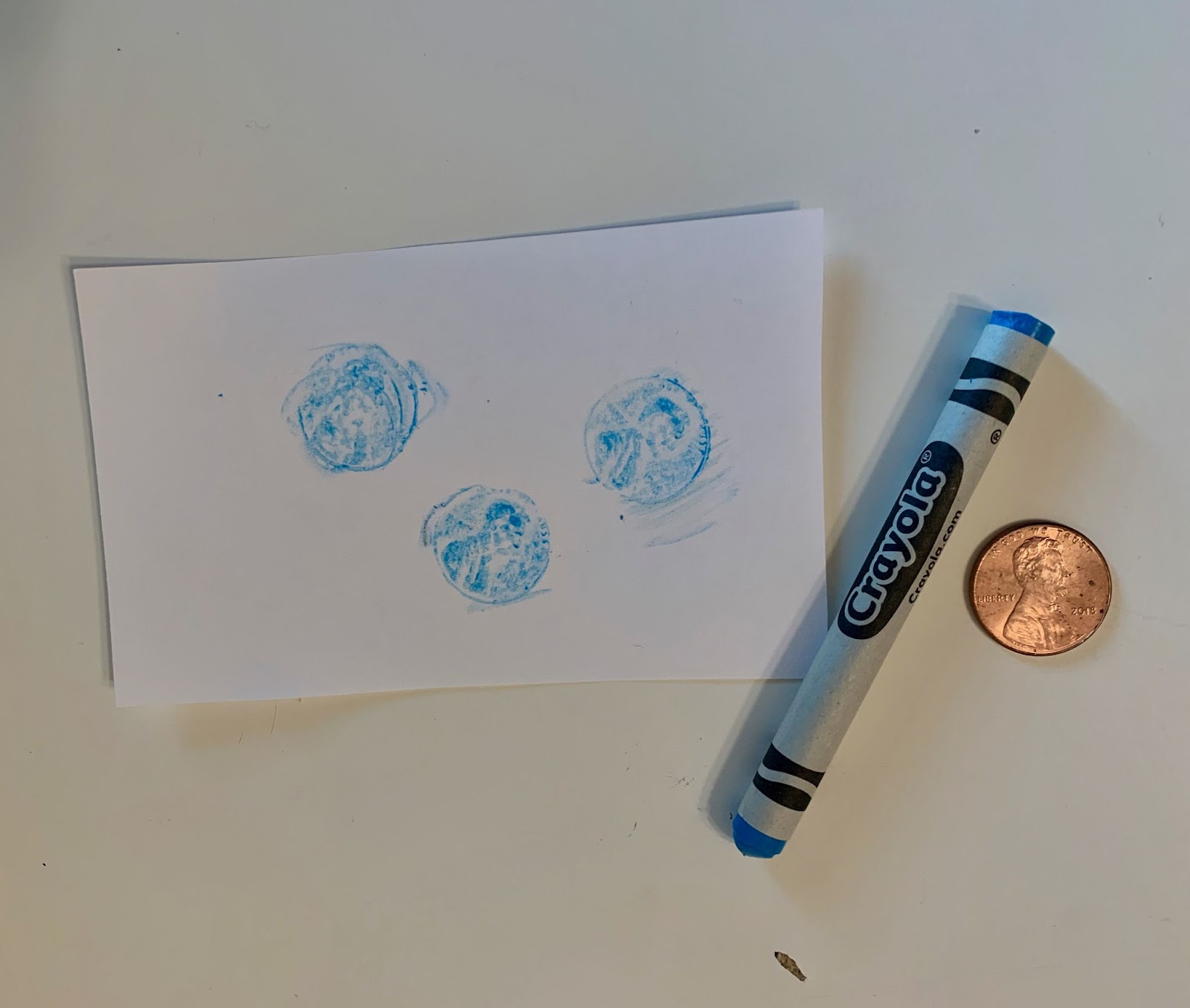 Penny rubbing art. Blue crayon and penny on a white surface with penny rubbings on paper.
