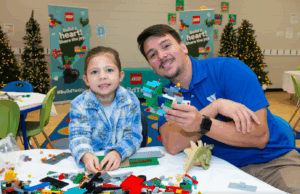 LEGO Build to Give event: Girl and adult building with LEGO bricks. Christmas trees in background. Holiday toy drive.