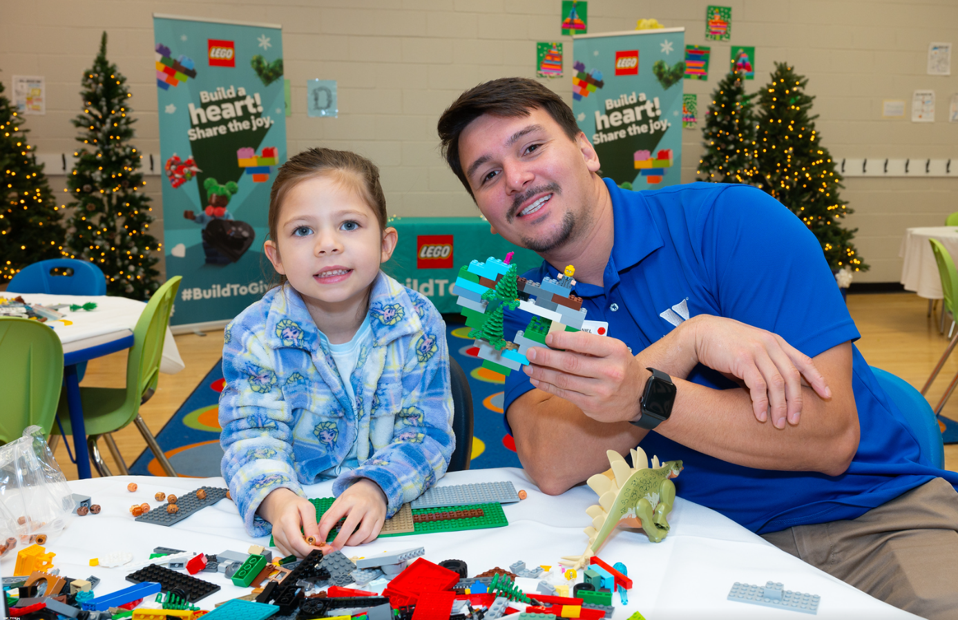 LEGO Build to Give event: Girl and adult building with LEGO bricks. Christmas trees in background. Holiday toy drive.