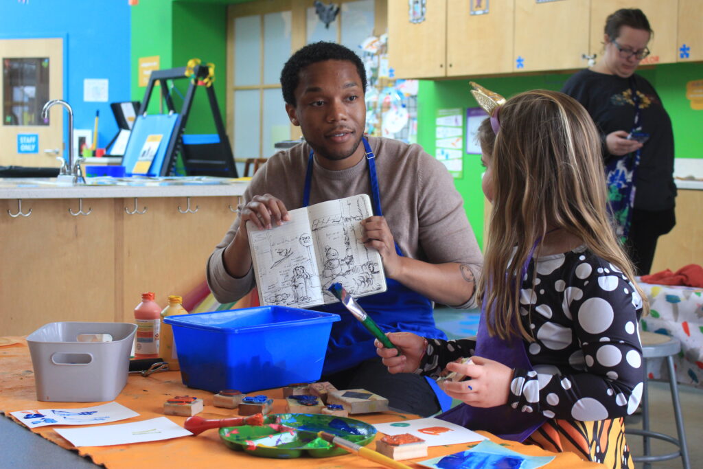 Creative in Residence: Man shows sketchbook to child during art session. Art supplies visible. IMG_6173.