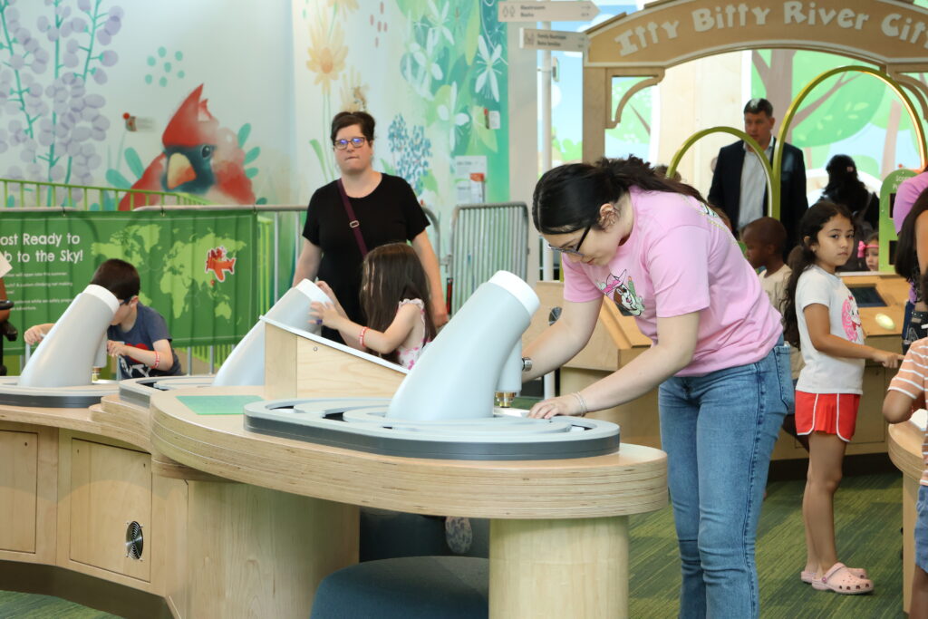 Teenager in a pink t-shirt examines a display at a curved wooden table alongside children using interactive scopes.