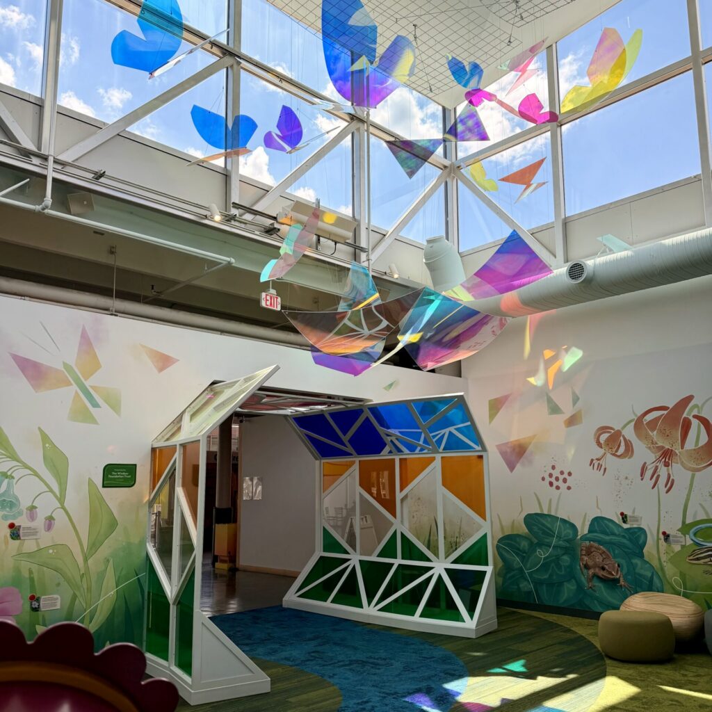 Colorful iridescent butterfly sculptures hang from the ceiling above a geometric tunnel in a bright, mural-filled room.