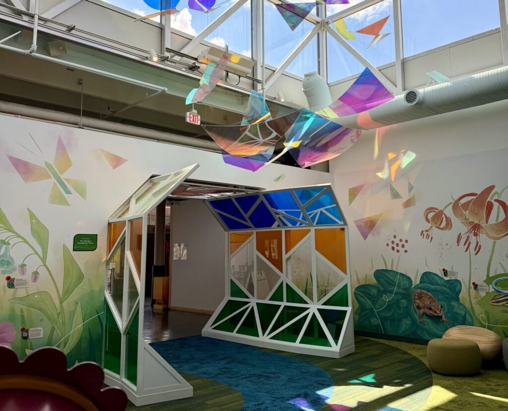 Colorful iridescent butterfly sculptures hang from the ceiling above a geometric tunnel in a bright, mural-filled room.