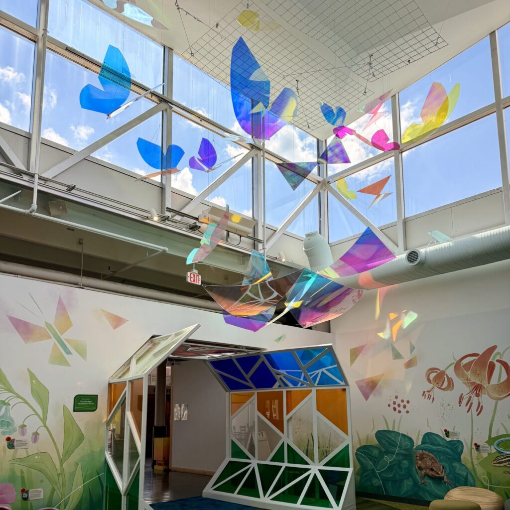 Colorful iridescent butterfly sculptures hang from the ceiling above a geometric tunnel in a bright, mural-filled room.