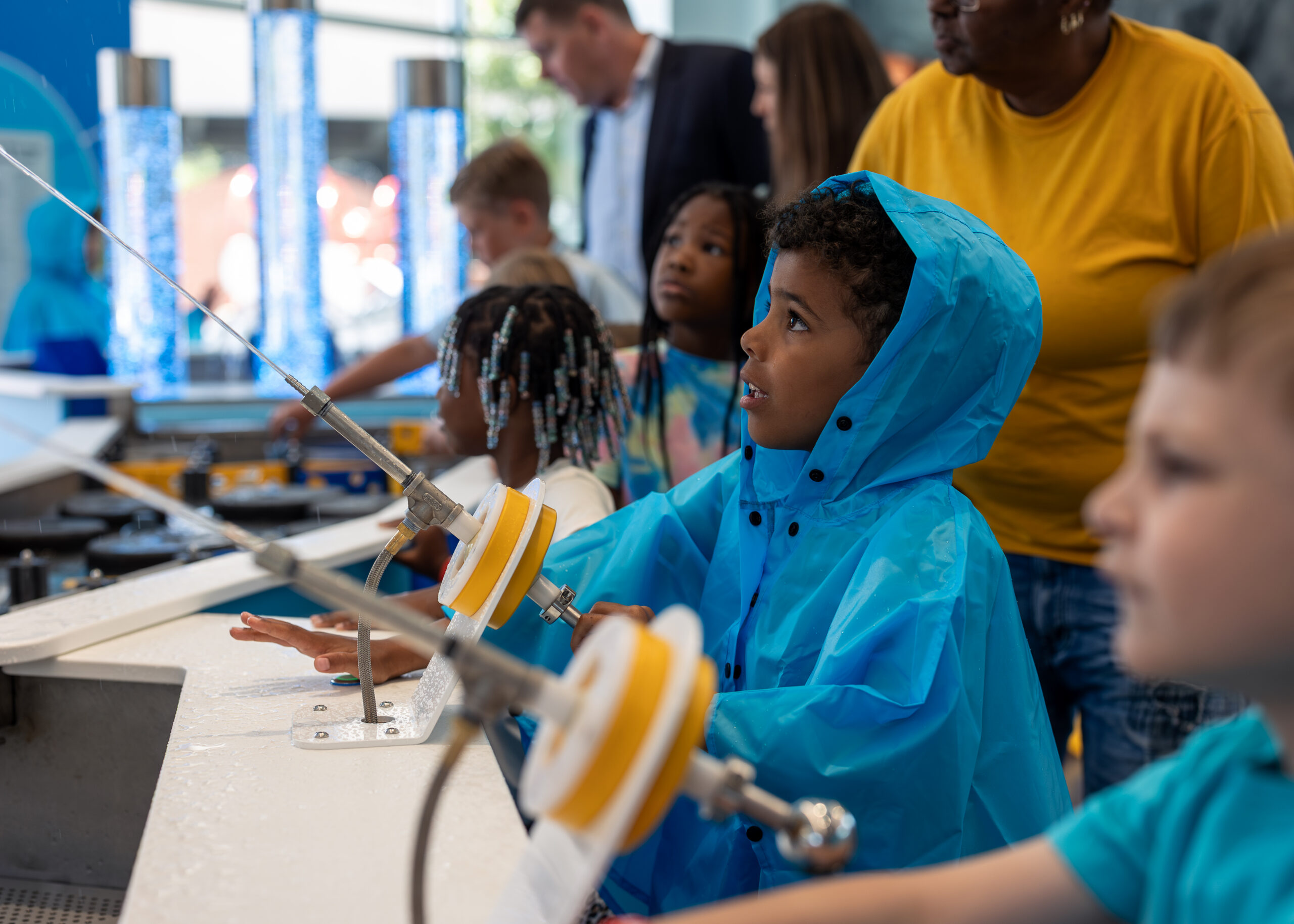 Boy in a bright blue hooded raincoat directs a high-pressure water stream at an interactive exhibit in a science center.