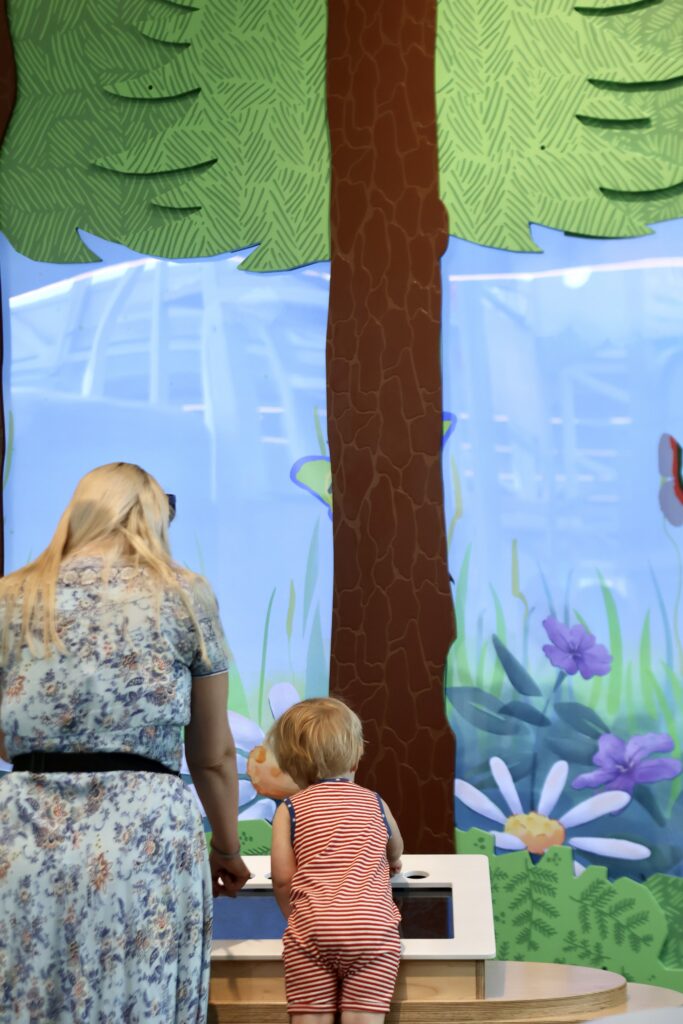 A woman and a toddler interact with an interactive nature-themed display featuring painted trees and wildflowers.