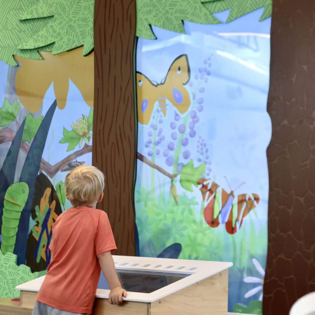 Blond child in an orange shirt stands before a colorful Nature Lab wall mural depicting butterflies and woodland trees.