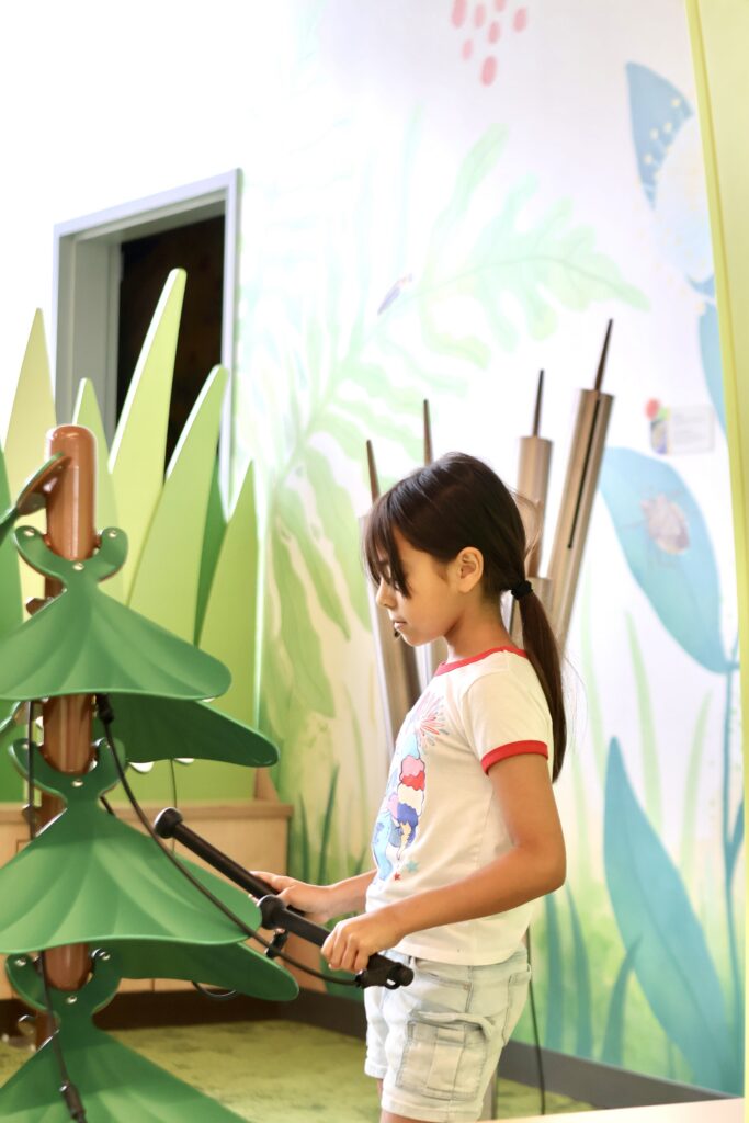 Young girl strikes a green bell-tree instrument in a play area decorated with botanical wall murals.