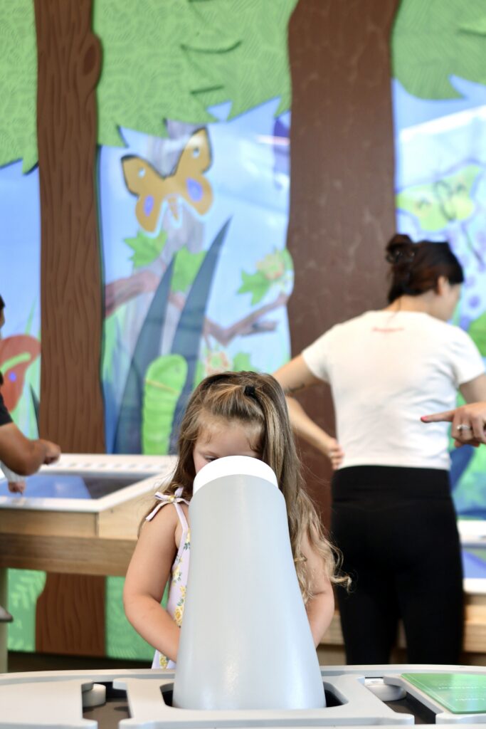 Young girl looks through a white viewing scope in a Nature Lab display featuring illustrated trees and butterflies.