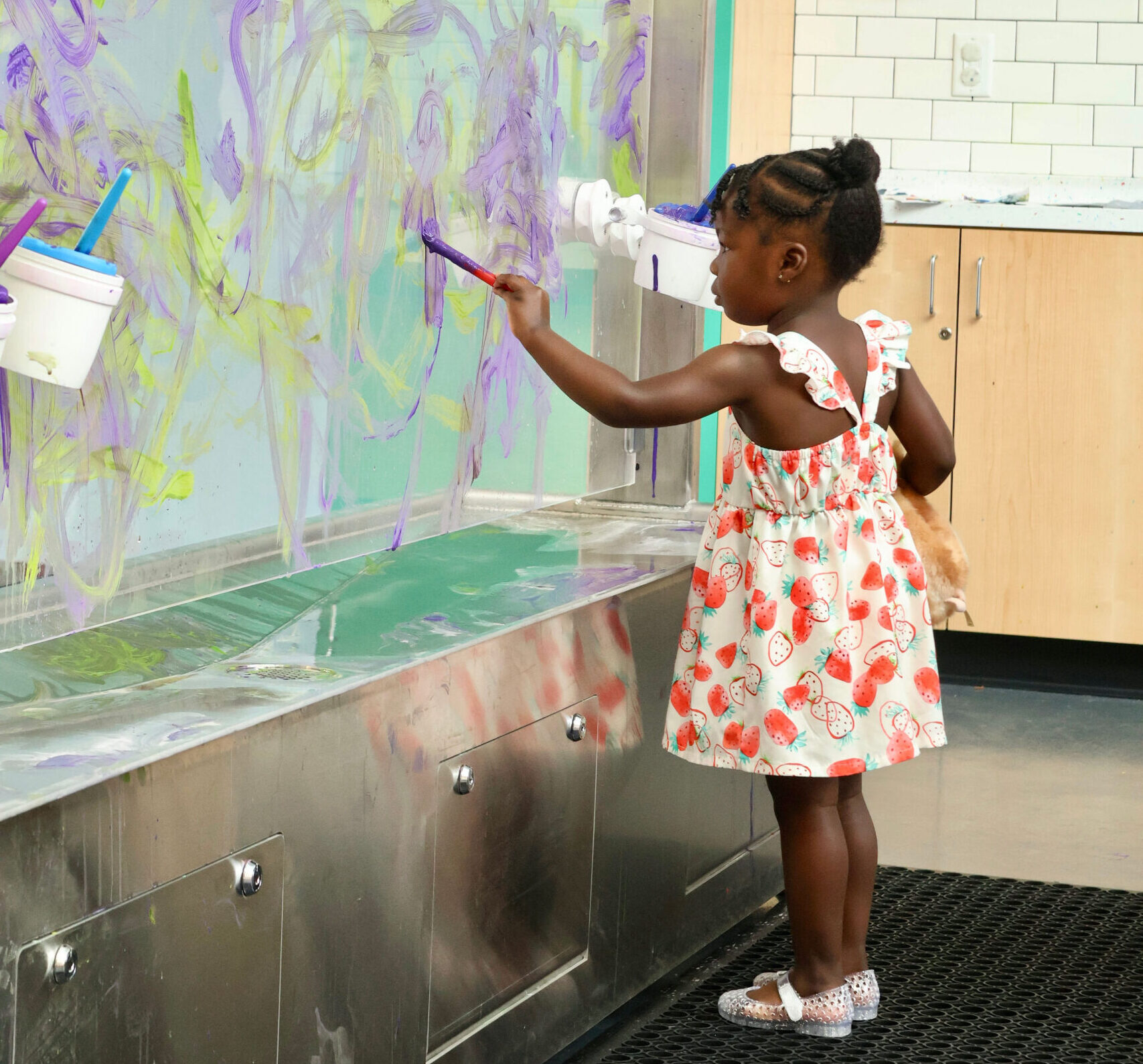 Young girl in a strawberry dress paints purple and green streaks on a large glass wall in Seymour Studio.
