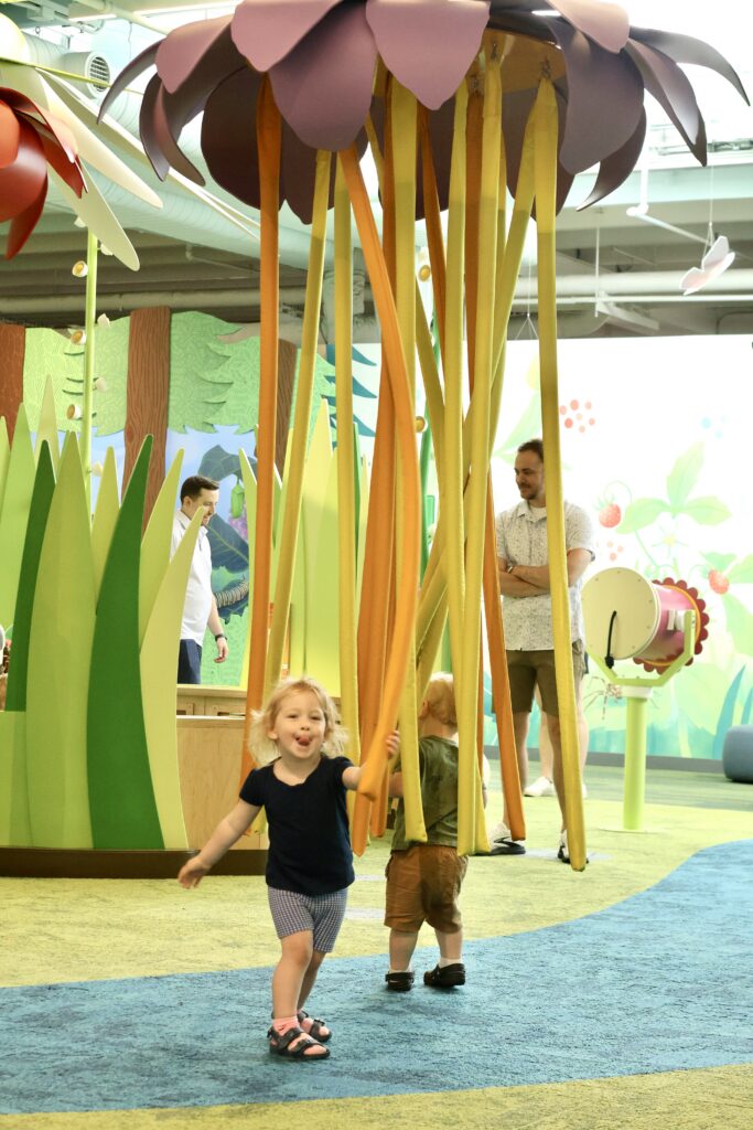 Toddler sticks out her tongue while walking under dangling golden ribbons hanging from a giant purple flower sculpture.