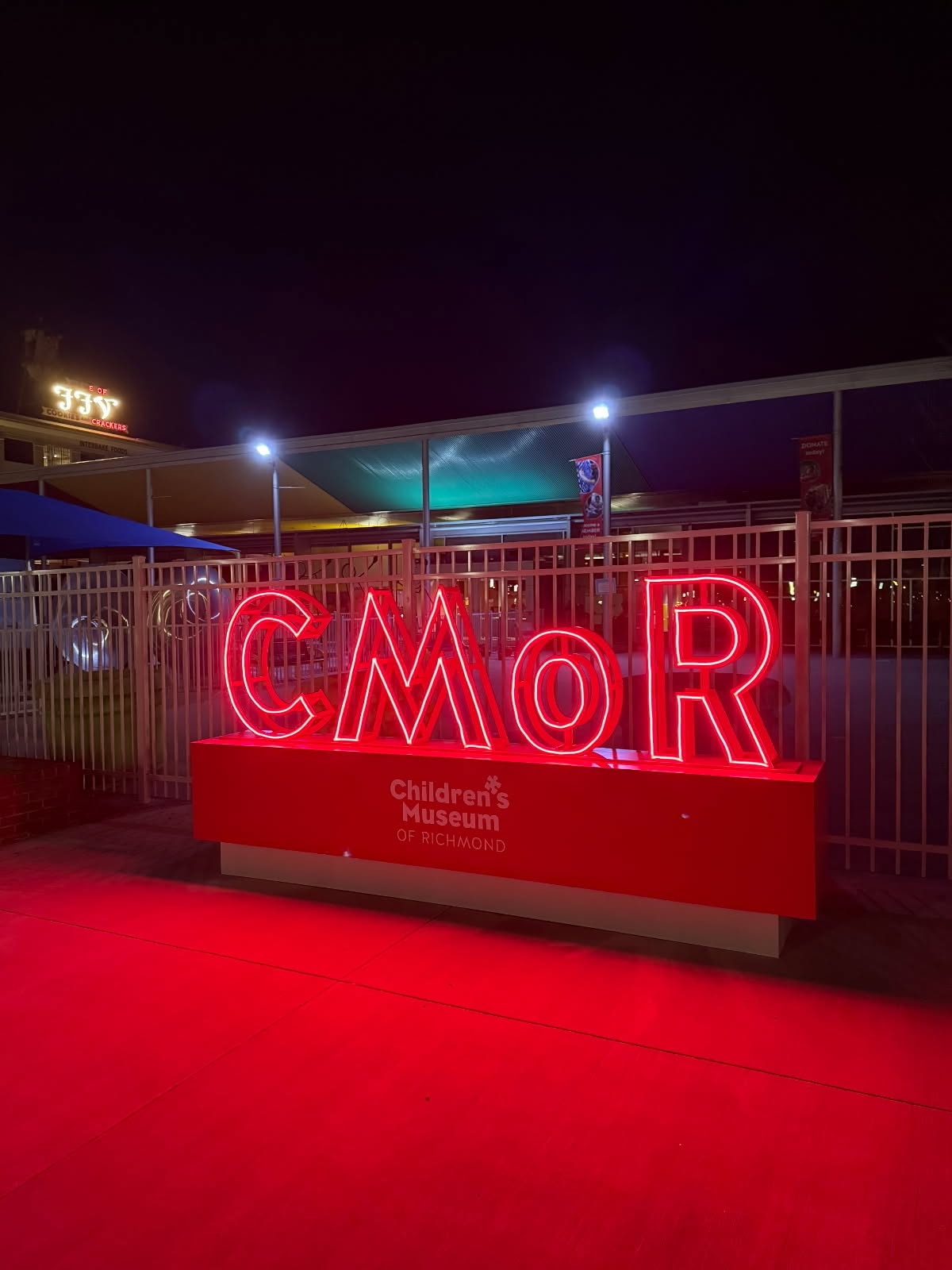 Bright red neon CMOR sign and Children's Museum of Richmond logo illuminated at night in front of a metal fence.