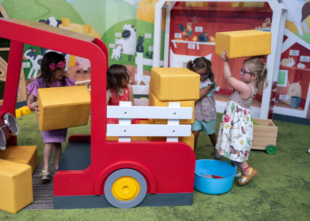 Four young children play with yellow foam blocks around a red toy truck in a farm-themed indoor playground.