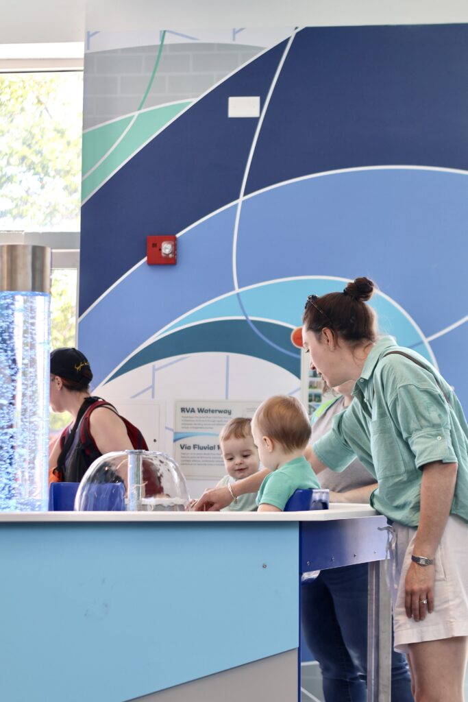 Mother and young children play at a water table in front of a blue geometric mural at the RVA Waterway exhibit.