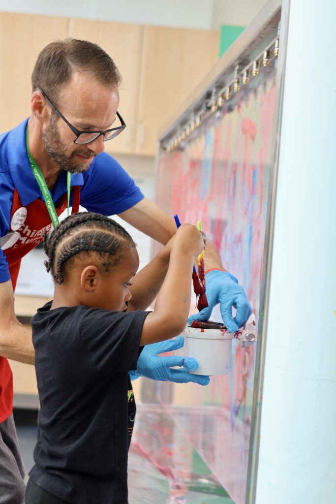 Man wearing a red Children's Museum apron helps a young child paint on a clear vertical surface with brushes and red paint.