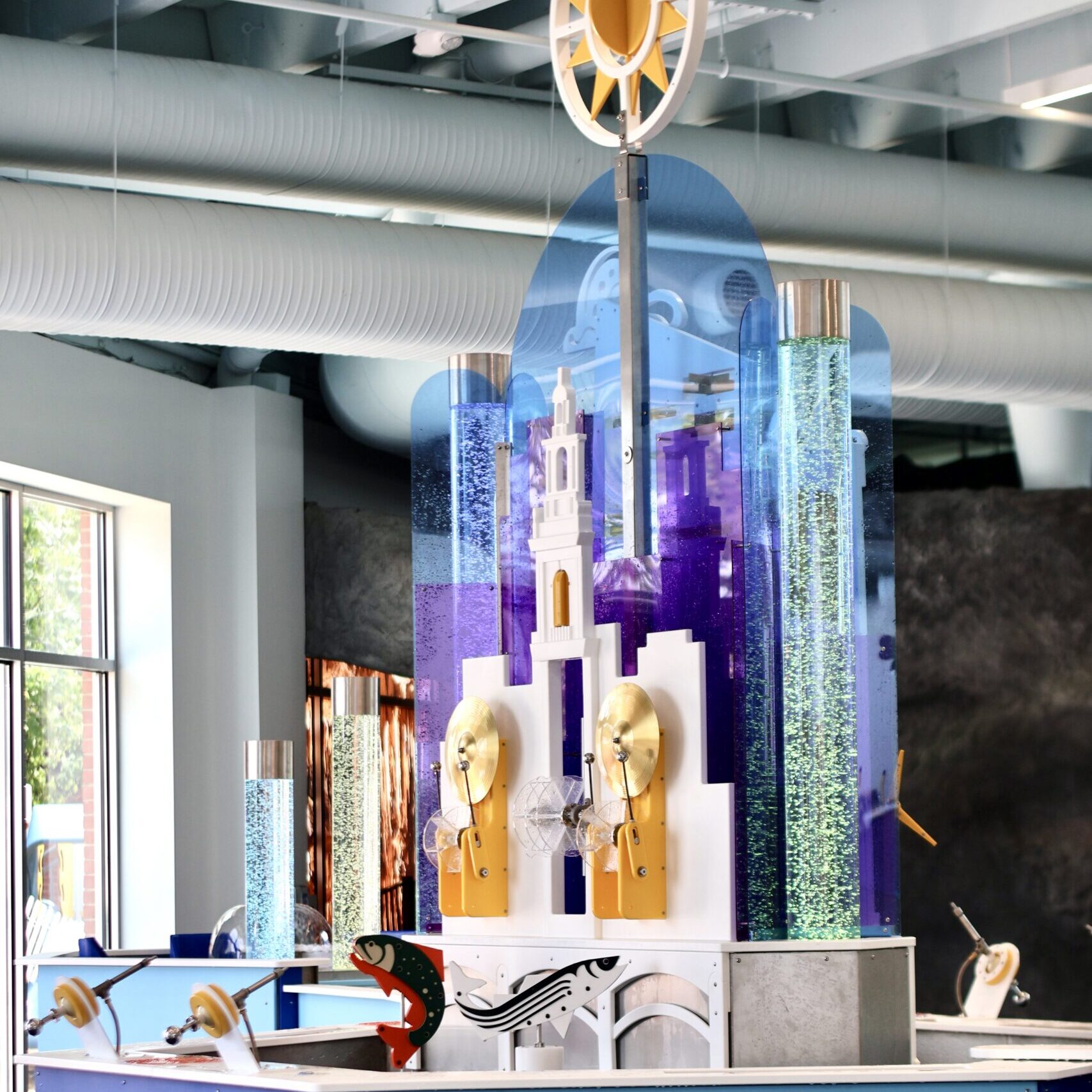 Interactive water exhibit with bubble tubes, spinning gold discs, and stylized fish models in a brightly lit room.