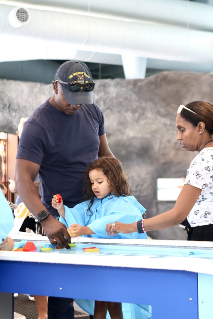 Father and mother helping their young daughter build with colorful toy blocks on a blue activity table.