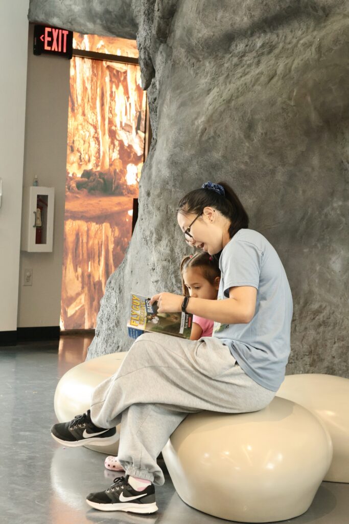 Young woman reading a Fly Guy Presents: Bats book to a young girl while sitting on a smooth bench near a cave display.