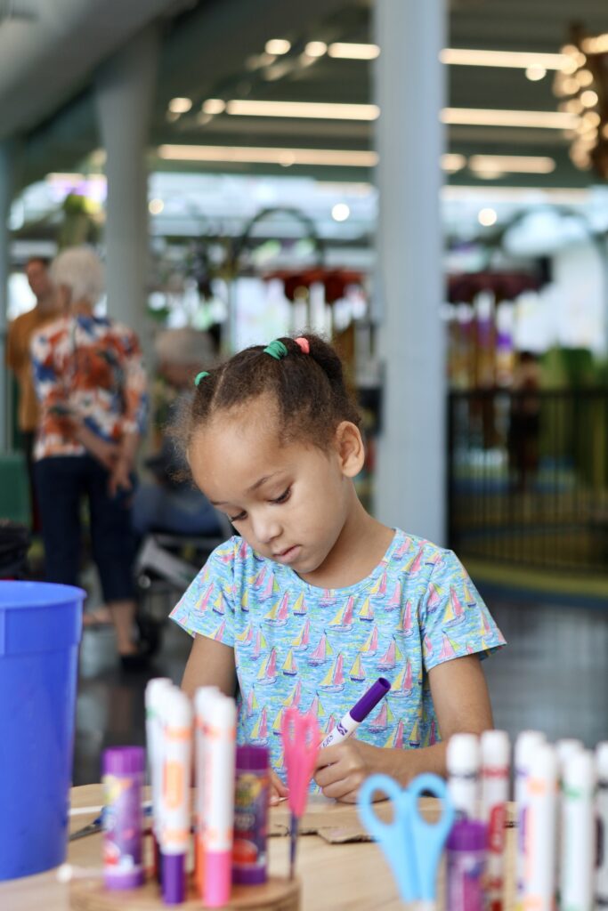 Young girl with hair in small buns drawing with a purple marker at a table filled with colorful art supplies.