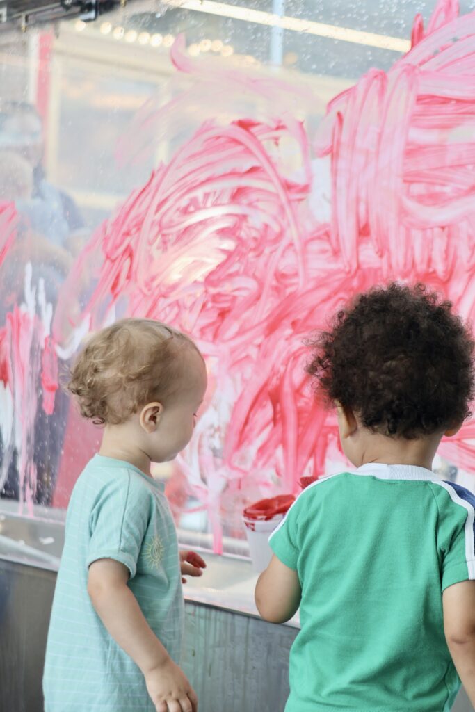 Two toddlers paint with red pigments on a clear glass window.