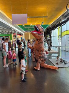 Children and adults watch an inflatable dinosaur costume standing in a museum near a large fossil display.