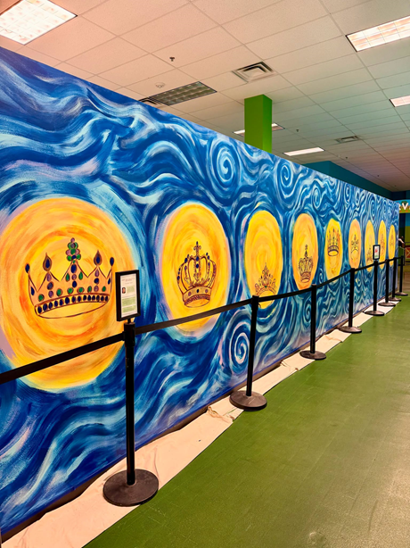 Large mural featuring painted golden crowns set against swirling blue and yellow patterns behind a black stanchion rope.