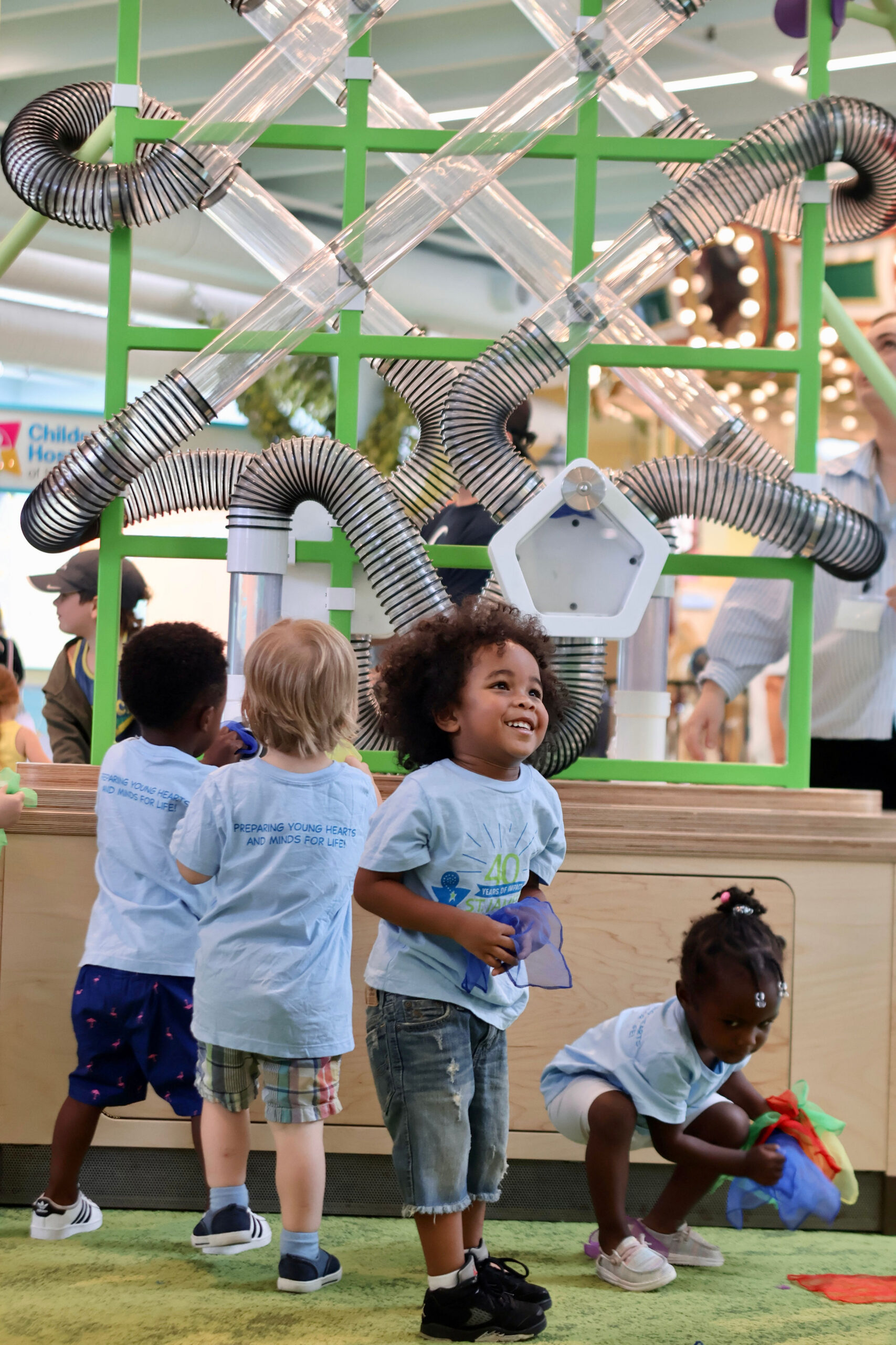 Children in light blue shirts interact with a green structure featuring interconnected clear plastic tubes.