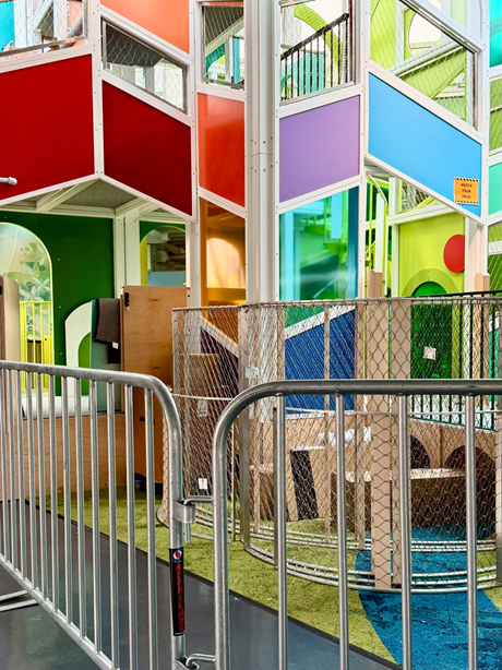 Multi-colored indoor Skybox climber playground structure behind metal safety fencing.