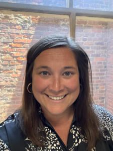 Smiling woman with long brown hair wearing silver hoop earrings and a black patterned top against a brick wall.