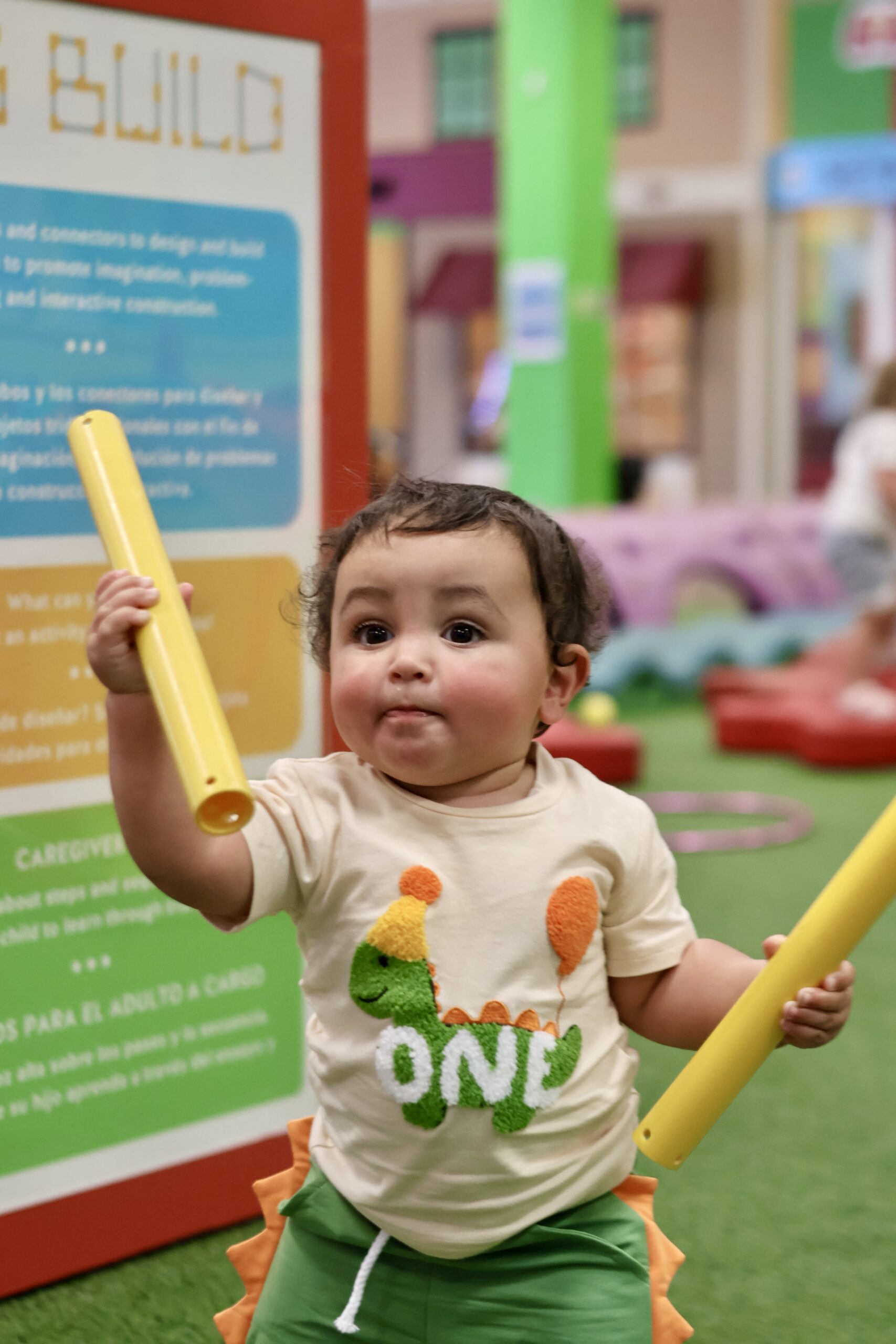 Toddler in dinosaur 'ONE' birthday shirt holds yellow tubes at an indoor play area.