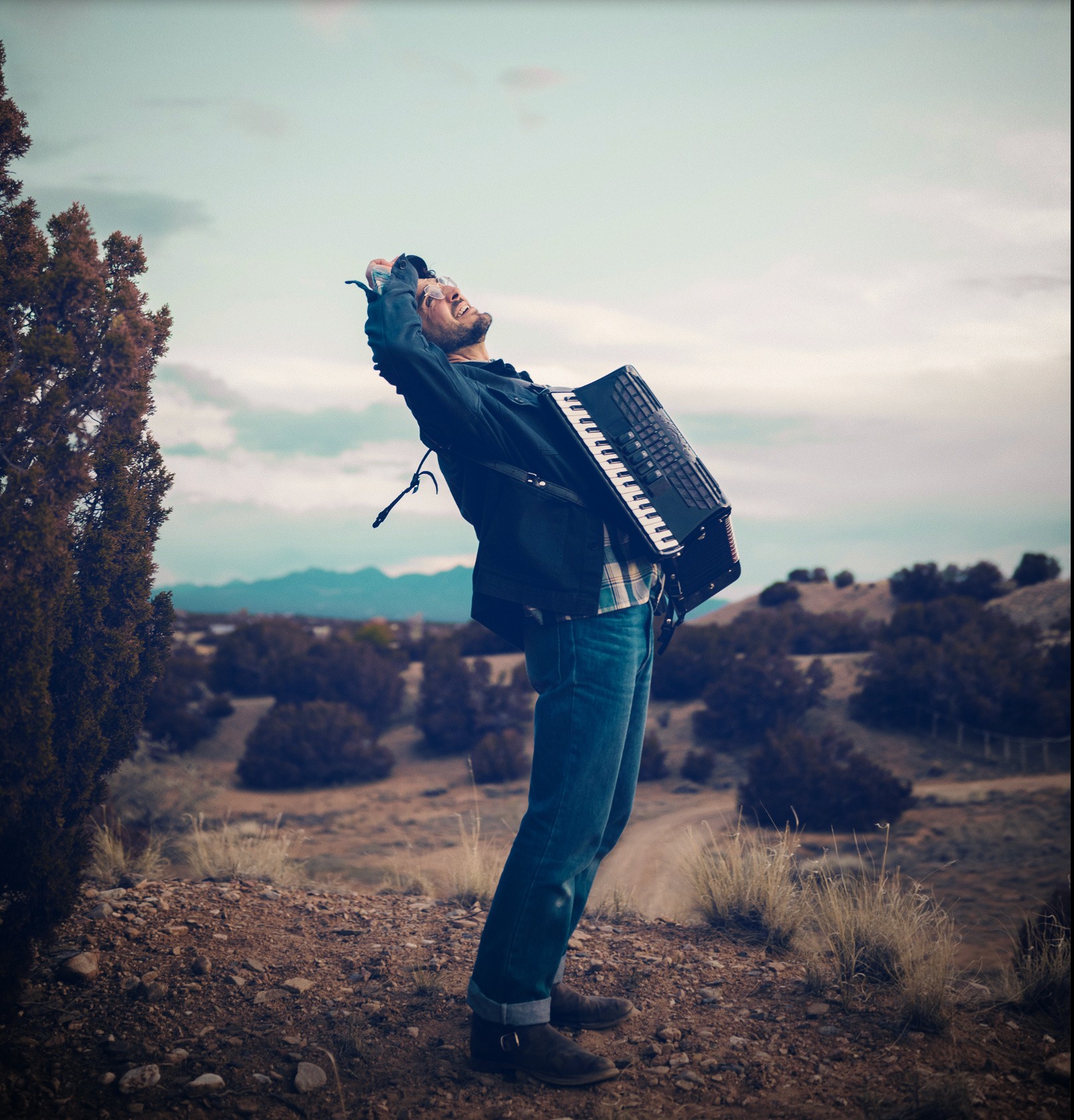 Man wearing a denim jacket leans backward while playing a black accordion on a desert hillside at twilight.