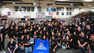 Large group of students in black robotics team shirts posing with a blue FIRST Robotics Competition banner in a gym.