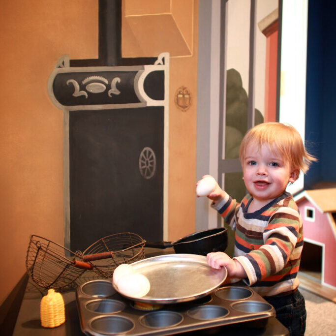 Framed: Step Into Art. A toddler plays with eggs in a toy kitchen, vintage oven, and barn in the background.