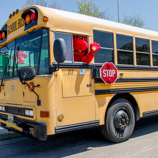 Mascot waving from a yellow school bus. Kindergarten readiness concept.