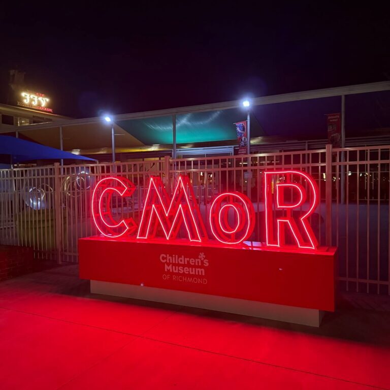 Bright red neon CMOR sign and Children's Museum of Richmond logo illuminated at night in front of a metal fence.