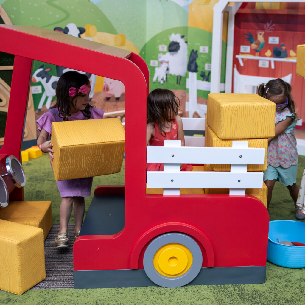 Four young children play with yellow foam blocks around a red toy truck in a farm-themed indoor playground.