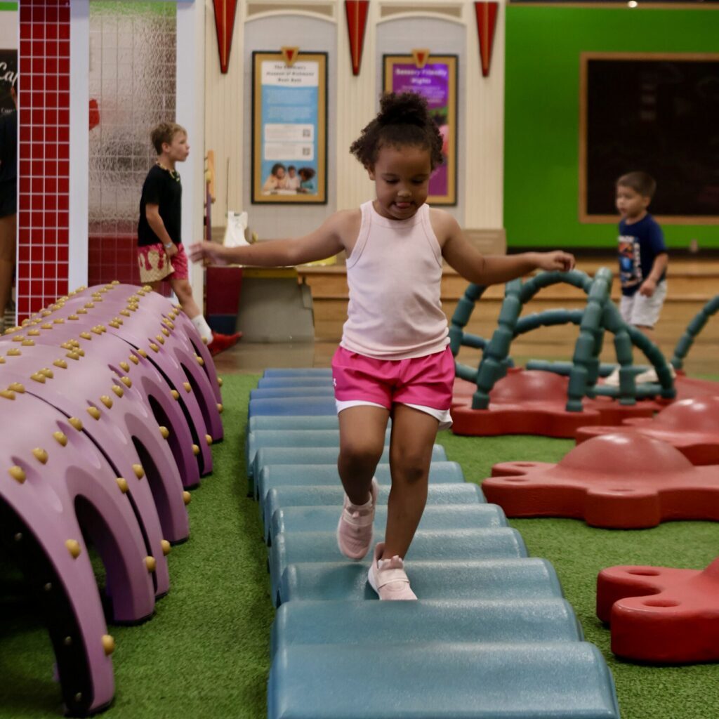 Young girl balances across a blue foam bridge in an indoor playground near a pizza cafe and children's theatre sign.