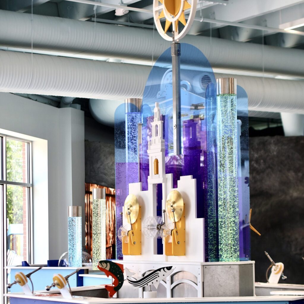 Interactive water exhibit with bubble tubes, spinning gold discs, and stylized fish models in a brightly lit room.