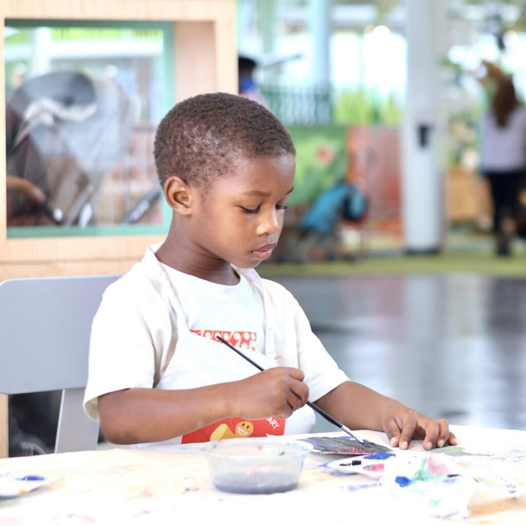 Young boy in a white t-shirt painting with a brush at a table in a brightly lit indoor space.