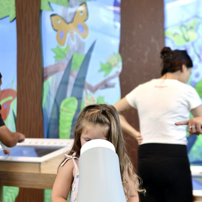 Young girl looks through a white viewing scope in a Nature Lab display featuring illustrated trees and butterflies.