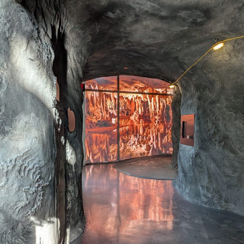 A simulated cave tunnel with textured gray walls leading to a brightly lit display of illuminated stalactites.