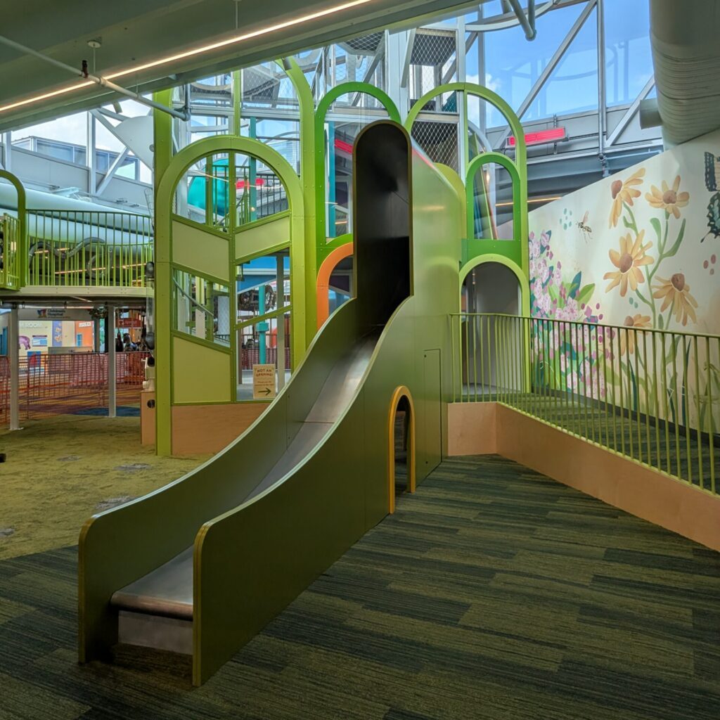 Indoor playground with a tall green slide and a wall mural featuring painted flowers and insects.