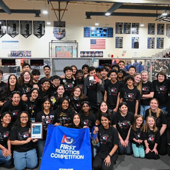 Large group of students in black robotics team shirts posing with a blue FIRST Robotics Competition banner in a gym.