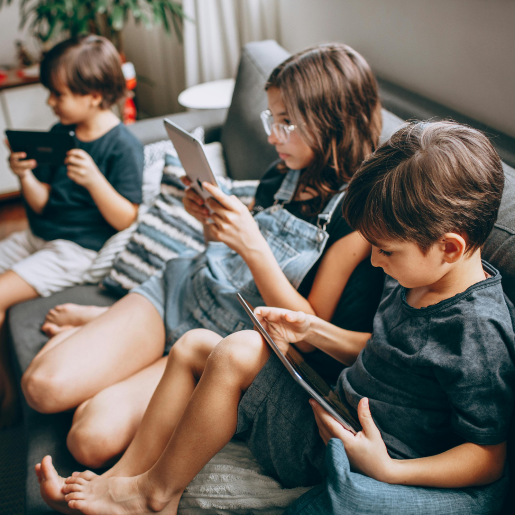 Three children sitting on a grey couch, each focused on using a tablet device.