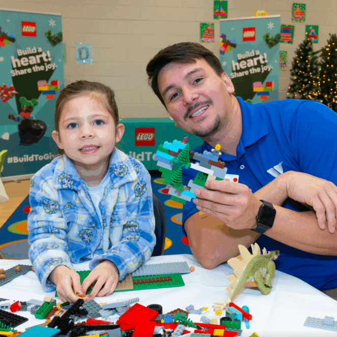 LEGO Build to Give event: Girl and adult building with LEGO bricks. Christmas trees in background. Holiday toy drive.