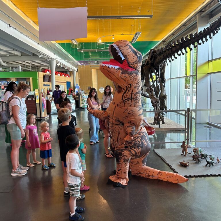 Children and adults watch an inflatable dinosaur costume standing in a museum near a large fossil display.